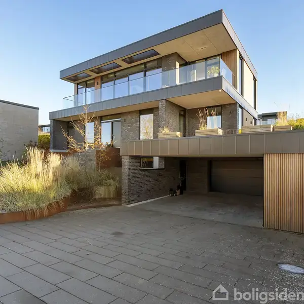 En moderne to-etagers bolig med store glasvægge og en balkon øverst, placeret i et stilfuldt boligområde. Foran huset ses en stor indkørsel og velplejede planter.