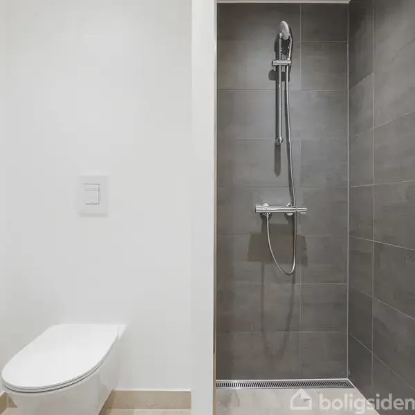 Toilet og bruser i et moderne badeværelse. Toilettet er væghængt til venstre, mens bruseren med håndbruser er placeret til højre på en mørk flisebelagt væg.