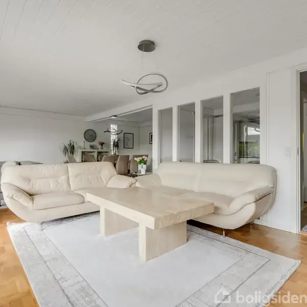 Et lyst opholdsstue med to cremefarvede sofaer omkring et firkantet træbord. Rummet har parketgulv, tæppe, og store vinduer, der tillader naturligt lys. Moderne indretning med planter og kunstværker.