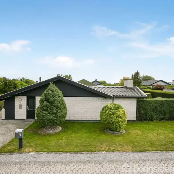Et enfamiliehus med fladt tag står midt i en velholdt have med to klippede buske. En brolagt indkørsel fører til hoveddøren. Hække omgiver ejendommen fra begge sider.