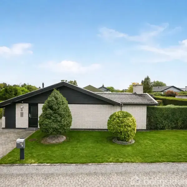 En enkel etplanshus med fladt tag står omgivet af en grøn have med trimmet græs og to buske. Huset har en brolagt indkørsel og ligger i et fredeligt boligområde.
