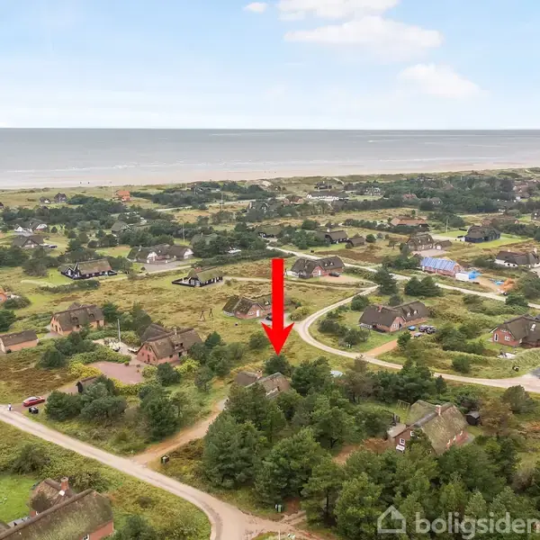 En rød pil peger mod et hus omgivet af træer i et kystnært sommerhusområde. Veje og flere huse er spredt i landskabet, mens stranden og havet ses i baggrunden.