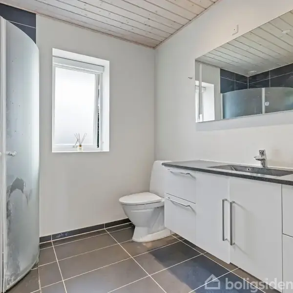 Badeværelse viser en brusekabine ved siden af et vindue. Til højre er et hvidt badeværelsesskab med vask, stort spejl og mørke gulvfliser.