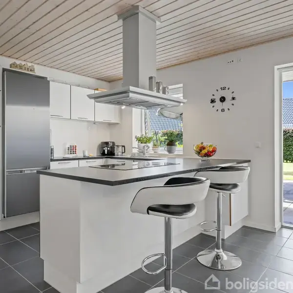 Et moderne køkken har en central ø med barstole, emhætte og frugt på bordet. Til venstre står hvide skabe og et sølv køleskab. Store vinduer åbner mod en have.