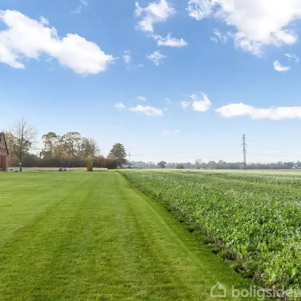 En stor grøn mark strækker sig mod horisonten under en klar blå himmel med få skyer. Til venstre står en rød murstensbygning omgivet af træer.