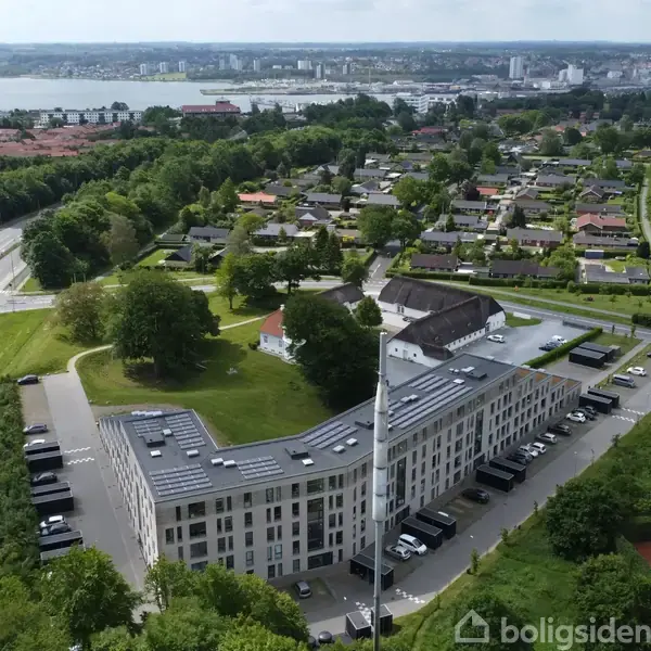 En stor, hvid bygning strækker sig langs en vej, omgivet af træer og parkeringspladser. I baggrunden ses en by og en sø, der skaber en smuk panoramaudsigt.