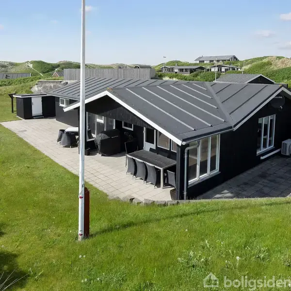 Sort sommerhus står på en græsplæne; fliser og terrassemøbler ses omkring huset. Baggrund viser klitter og andre huse under en blå himmel.