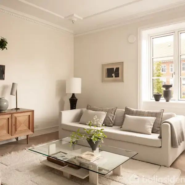 En lys stue med en hvid sofa fyldt med puder, placeret ved et vindue. I rummet er der også et glasbord, grønne planter og en trækommode.