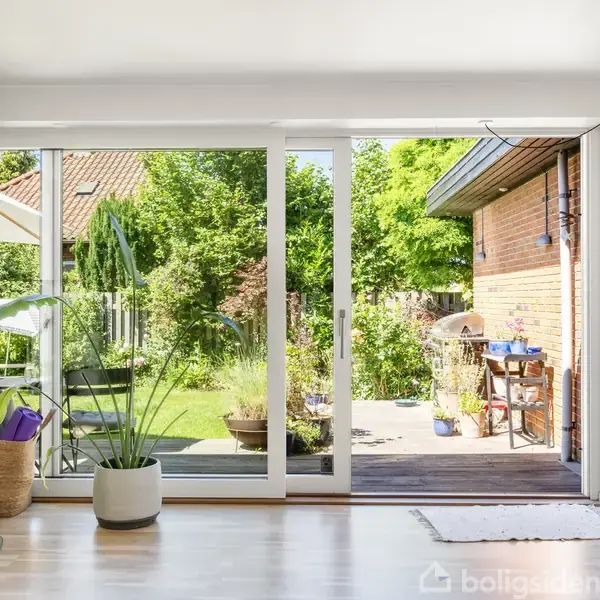 Stuen forbindes med haven gennem en stor skydedør. En grøn plante står i en potte ved døren. Udenfor er en terrasse og frodig vegetation.