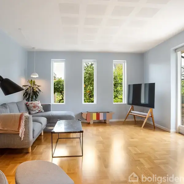 En stue med en grå sofa, der står ved siden af en gulvlampe. Rummet har lyseblå vægge, parketgulv og tre smalle vinduer. En fladskærms-TV står på et stativ.