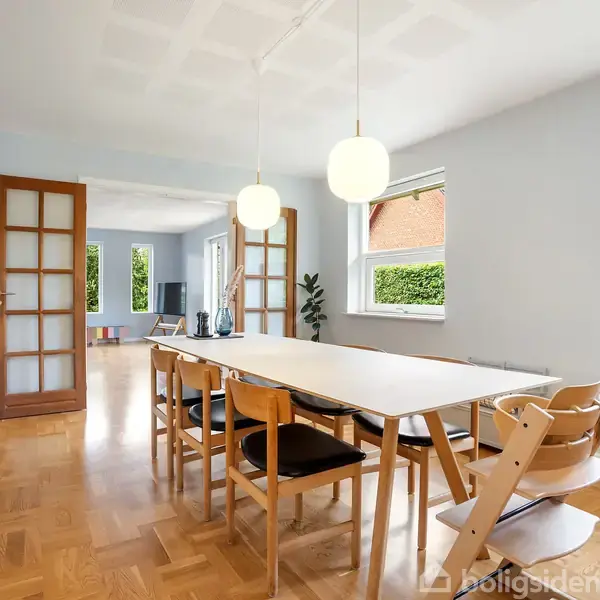 Langbord omgivet af stole står i et lyst rum med parketgulv. To hængelamper er tændt over bordet. Store glasdøre fører til yderligere rum, og vegetation ses udenfor vinduerne.