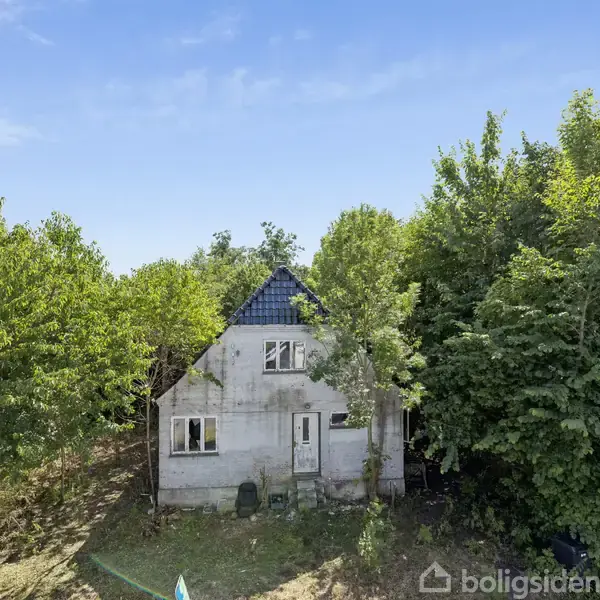 Et gråt hus med blåttegl tag står stille omgivet af tætbevokset skov. Sollys filtrerer ned gennem træerne og kaster skygger på husets facade.