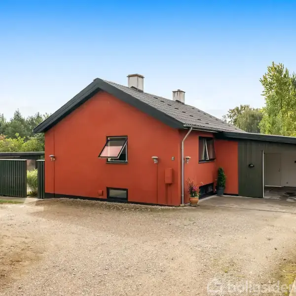 En lille træbygning står på en flisebelagt terrasse, omgivet af grønne buske og græsplæne. Bygningen har store vinduer, og foran står en sofa med et bord.