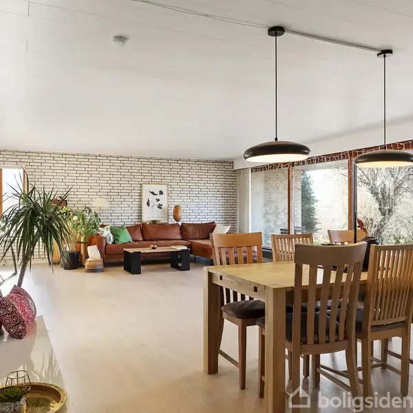 Stue med træbord, seks stole, og hængende lamper. Brun lædersofa står foran en hvid murstensvæg. Store vinduer giver udsigt til have. Planter og kunstværk tilføjer dekoration.