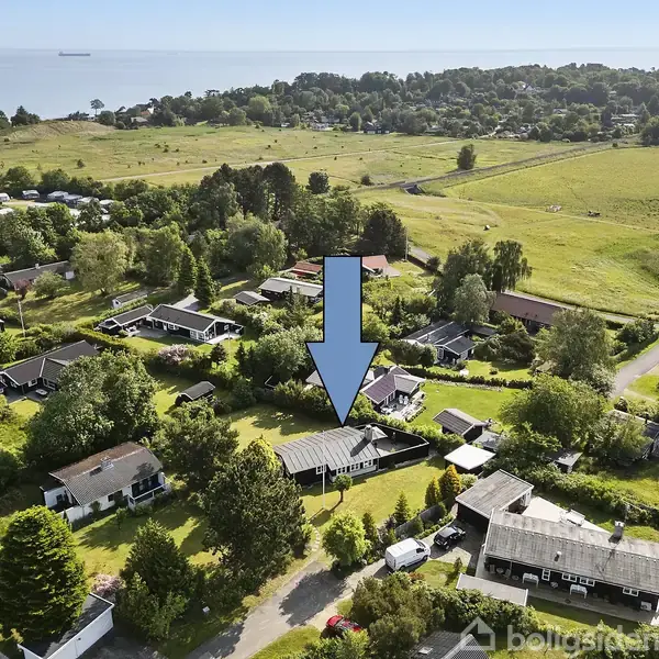 En blå pil peger på et hus omgivet af træer og andre huse; landskabet består af grønne marker og en kystlinje i baggrunden.