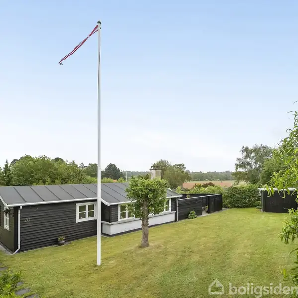 Et sort sommerhus står på en velplejet græsplæne, omgivet af grønne træer. En høj flagstang hejser et flag. Baggrunden viser åbne marker og en klar blå himmel.
