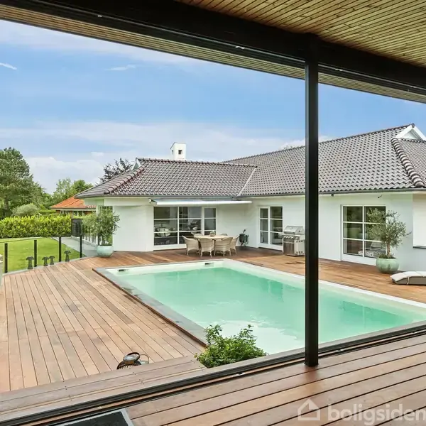 Hus med hvid facade og mørkt tag. Træterrasse med swimmingpool i midten. Grønt haveområde rundt omkring. Terrassebord og stole ved siden af poolen.