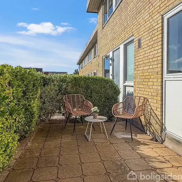 To flettet stole og et lille bord med blomster står på en flisebelagt terrasse foran en gul murstensbygning, omgivet af grønne buske under en blå himmel.