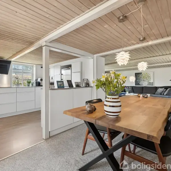 Bord med stribede vase, blomster og koppe, står i en stue med åbent køkken. Træloft og moderne møbler præger rummet, med loftslamper og sofa i baggrunden.