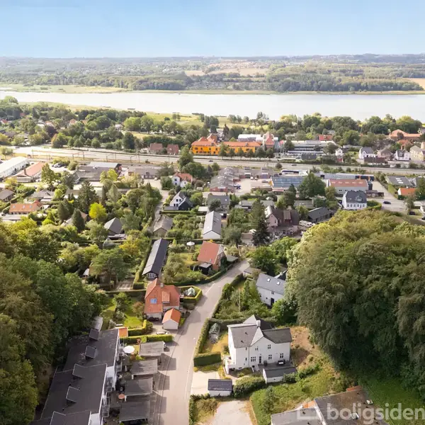 Luftfoto viser en bydel med snoede veje og spredte huse omgivet af tætte træer. I baggrunden ses en flod, og området er rigt på grønne områder og skovlandskab.