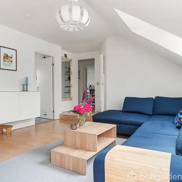 En blå hjørnesofa står i et moderne stuehjørne med skråloft. Et lavt bord foran sofaen bærer en blomsterbuket. Væggene har kunstværker, og der er en lampe i loftet.