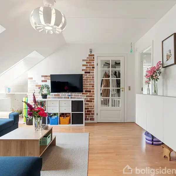 Stue med blå sofa foran et lavt sofabord; en væghængt fladskærm på murstensvæg bagved. Rummet har hvidt loft, skråvinduer og lys træparketgulv. Hylder og billeder på væggen.