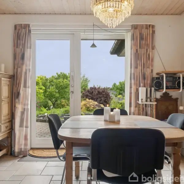 En spisebord med fire stole står i et lyst spisestueområde. En stor glasdør åbner ud til en have med frodige grønne planter. En elegant lysekrone hænger over bordet.