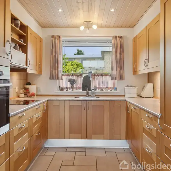 Køkken med træskabe, indbyggede ovne og keramisk komfur. Vindue over køkkenvask viser grønt udendørsområde. Fliser på gulvet, træloft med indbyggede spotlights.