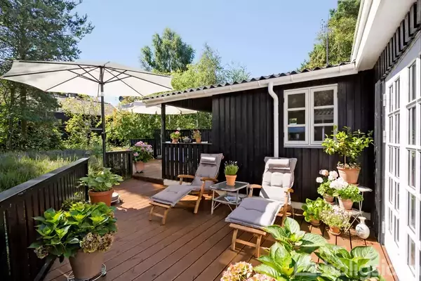 To liggestole står på en træterrasse omgivet af frodige planter. En stor parasol skygger for solen, mens sorte træfacader danner baggrund.