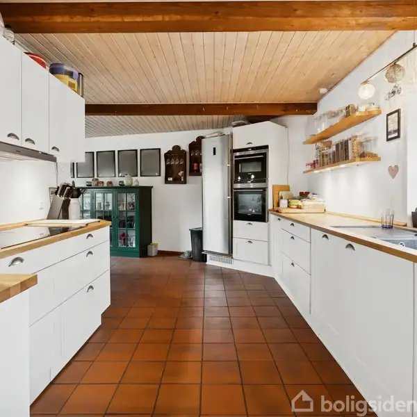 Et køkken med hvide skabe og træbordplader strækker sig langs begge vægge. Der er en ovn og skabe i baggrunden. Gulvet er dækket af terracottafliser. En enkeltvask er ved vinduet.