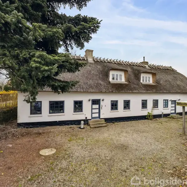 Bindingsværkshus med stråtag står i en gårdsplads omgivet af træer og buske. Huset har blå vinduesrammer og døre med en lille trappe ved indgangen.
