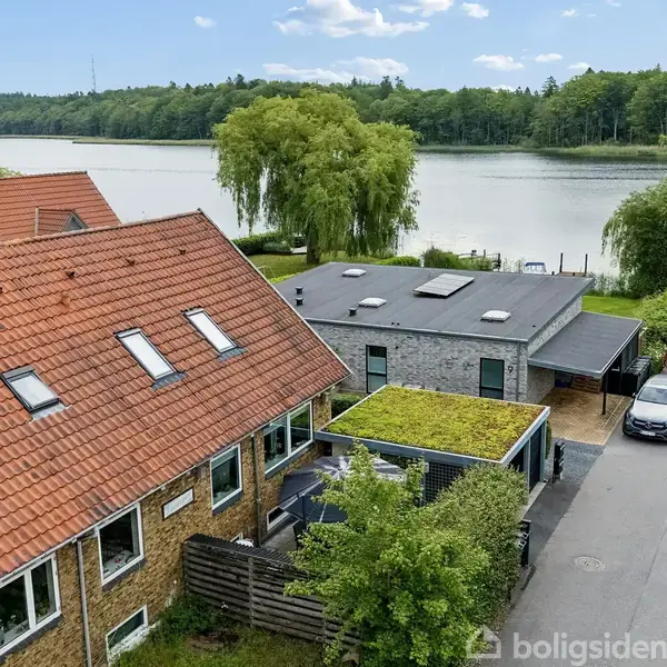 Et murstenshus med rødt tegltag og flere vinduer står ved en sø. En flad, moderne bygning ligger ved siden af, omgivet af grønne træer og parkerede biler.