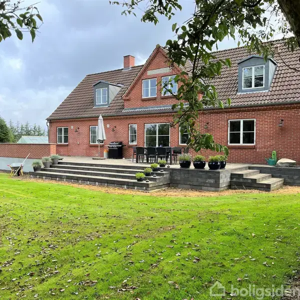 Et rødt murstenshus med brune tagsten står i fokus. Foran huset er en terrasse med havemøbler. Omgivelserne inkluderer en velplejet græsplæne og skov i baggrunden.