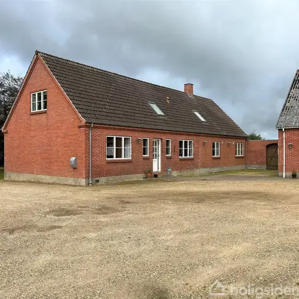 Et rødt murstenshus med hvide vinduer og dør står i en grusbelagt gårdsplads. Til højre ses et tilstødende bygning med et skifertag. Baggrund med træer og overskyet himmel.