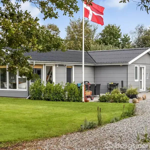 Gråt hus står stille i en velholdt have med grøn græsplæne, omgivet af buske. Det danske flag vajer fra en flagstang, og en grusvej fører til hoveddøren.