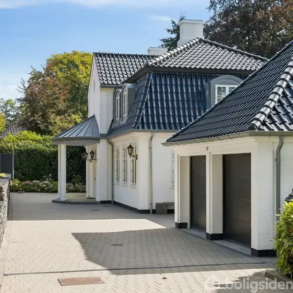 Et hvidt moderne hus står ved siden af en velplejet have med en glaspavillon. Haven er omgivet af grønne buske og træer under en klar blå himmel.
