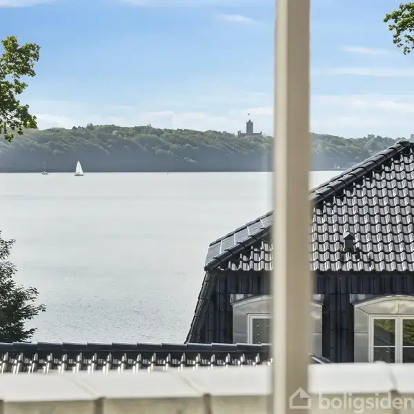 Et hvidt to-etagers hus med sort tegltag står stille i en velplejet indkørsel, flankeret af hække og træer, der skaber en privat og rolig atmosfære.