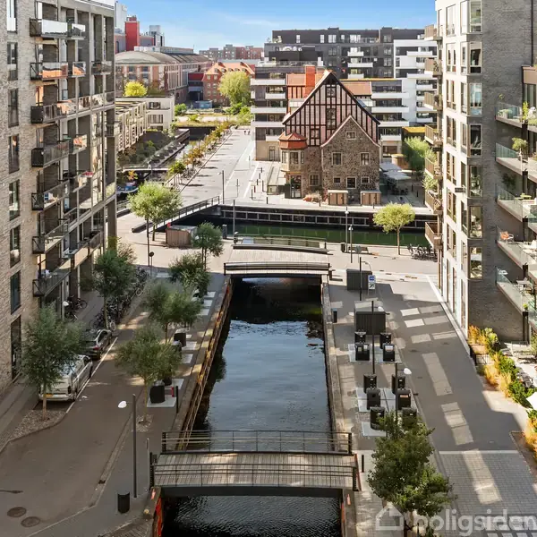 En bykanal løber gennem moderne boligbyggeri med altaner. En bro krydser kanalen, og i baggrunden ses et ældre hus med gavl. Sollys skinner på vejen.