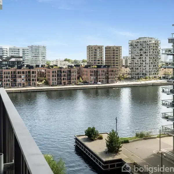 Balkonen har en glasrækværk og er møbleret med planter, der står stille. Den har udsigt over en bred kanal, med moderne lejlighedskomplekser på begge sider under en klar himmel.