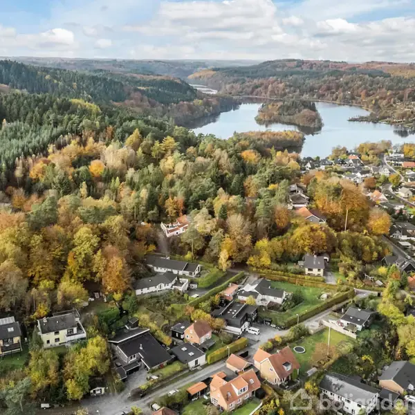 Et boligområde ligger ved en sø omgivet af skovklædte bakker. Huse er spredt blandt træerne, og veje snor sig gennem landskabet i efterårsfarver. I baggrunden ses en stor sø.