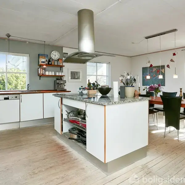 Et moderne køkken med en central køkkenø, der har en bordplade i granit. Køkkenet har hvide skabe og apparater. Der er et spisebord ved vinduerne med udsigt til en have.