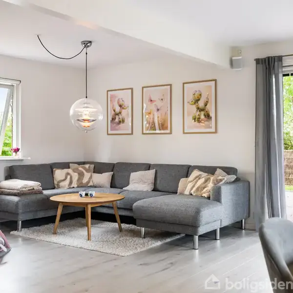 Grå hjørnesofa står i stue med lyse vægge. Over sofaen hænger tre billeder og en rund lampe. Et sofabord står foran, og et baby-legegymnastik er til venstre.