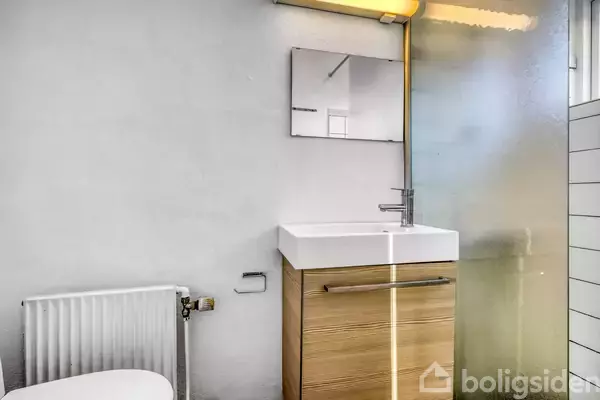 En moderne håndvask står ved en væg, mens en vandhane løber. Spejlet over vasken reflekterer rummet, som er lyst og har en minimalistisk stil med en brusevæg.