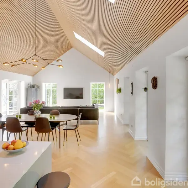 Et moderne, lyst køkken-alrum med spisebord, stole og sofa i midten. Loftet har trælameller og to ovenlysvinduer. Et vindue og dør på siden giver udsigt til en grøn have.