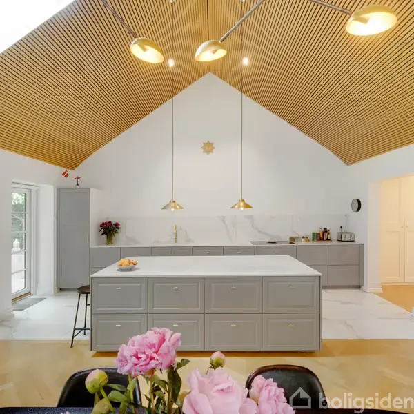 Åben køkkenområde med gråt køkkenø, tre hængende lamper og loft af trælameller. Lyserøde blomster står i forgrunden, mens hvide vægge skaber et lyst miljø.