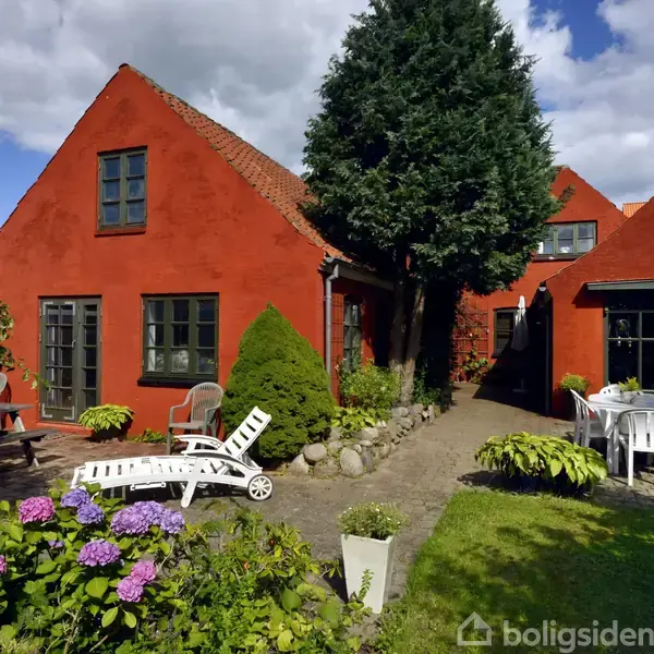 Rødt hus med hvide havemøbler i gårdhave. Blomsterbede med lilla hortensiaer omgiver området. Store træer kaster skygger på græsplænen under en blå himmel med skyer.