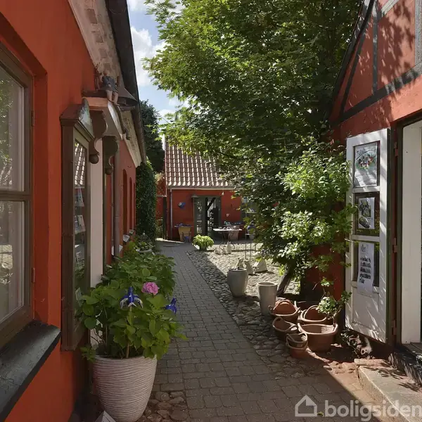 En smal gårdhave med røde murstensbygninger, hvor krukker med planter står i en række. Dør til højre åbner til en butik med postkort. Sollys gennemtrænger træernes blade.