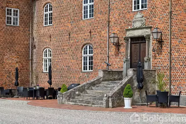 En gammel, muret bygning med buede vinduer har en stentrappe, der fører til en massiv trædør. Omkring trappen står sorte borde og stole, nogle med sorte parasoller.