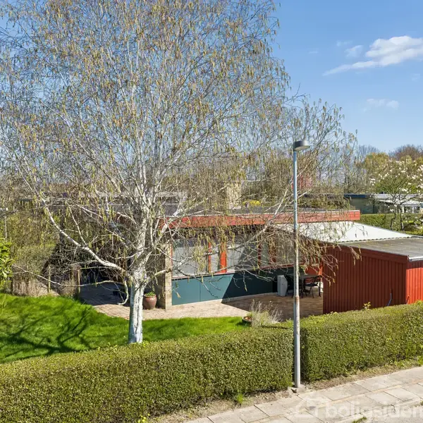 Et rødt hus ligger bag en hæk omgivet af have, med et stort træ og en lygtepæl i forgrunden. Himlen er klar med få skyer.