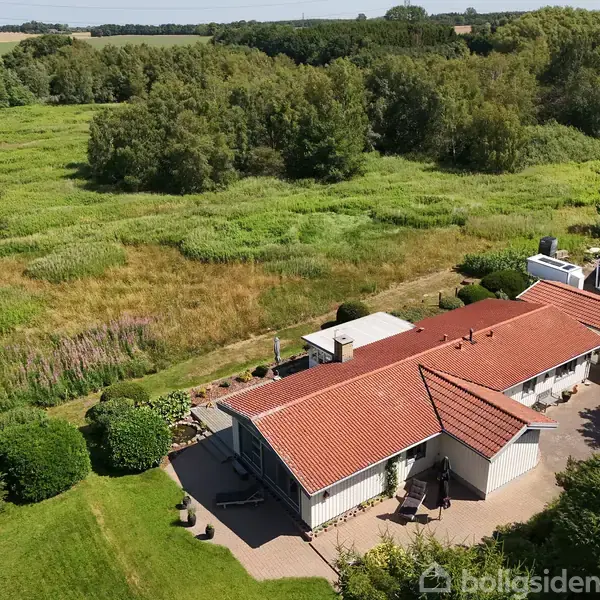 Hus med rødt tegltag; står stille; omgivet af frodig, grøn vegetation og græsareal ved siden af skov.
